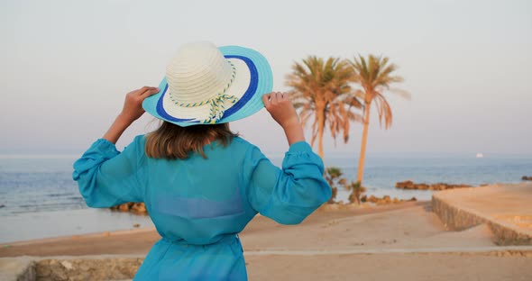 Teen Girl in Hat and Blue Dress on Tropical Beach Looking at Sea Rear View