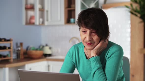 Elderly Woman Laughing and Smiling Using Laptop at Home in Kitchen alt