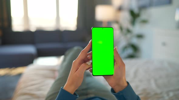 Woman at Home Lying on a Bed and Using Smartphone with Green Mockup Screen in Vertical Mode alt