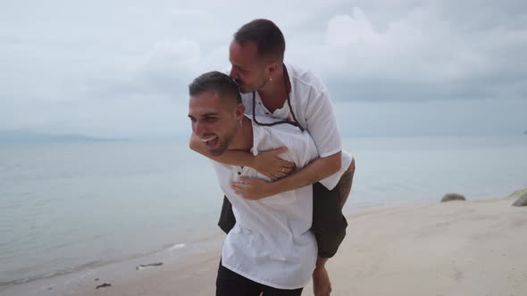 Cheerful Gay Man Giving Piggyback Ride to His Partner on a Beach alt