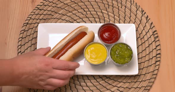 Top down view of a hand placing hot dogs on a plate beside ketchup, mustard, and relish. alt
