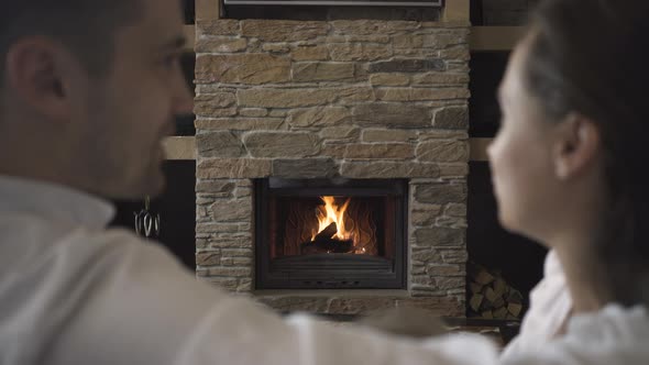 A man and a woman are talking in front of a fireplace with a fire burning in it alt