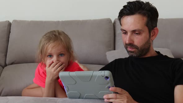Father and Daughter Sitting on Grey Sofa and Using Digital Tablet