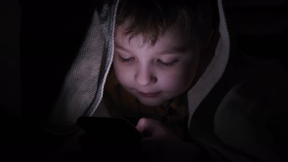 Little Cute Boy Sits at Night with a Phone Under the Blanket