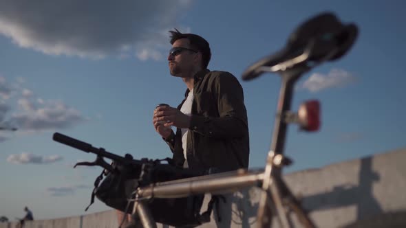 Male Cyclist Eats Sandwich and Drinks Coffee to Go While Sitting on Promenade By Sea alt