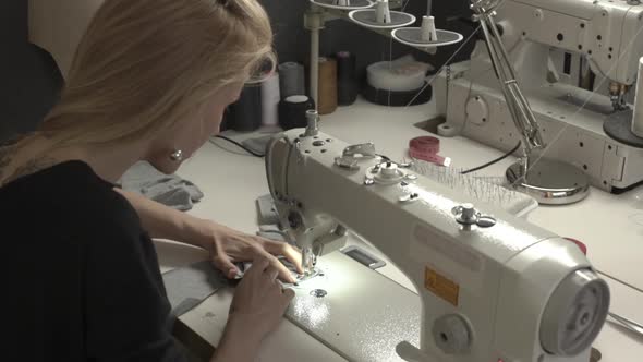 Blonde Caucasian Female in Black Dress Sitting at Table with Machine in Sewing Classroom Stitching alt