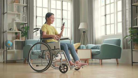 Asian Kid Girl Sitting In A Wheelchair While Video Calling Online In Mobile Chat App At Home alt