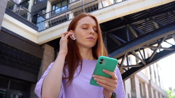 Joyful Pretty Redhead with Closed Eyes Listening Music Using Wireless Headphones in City Street alt