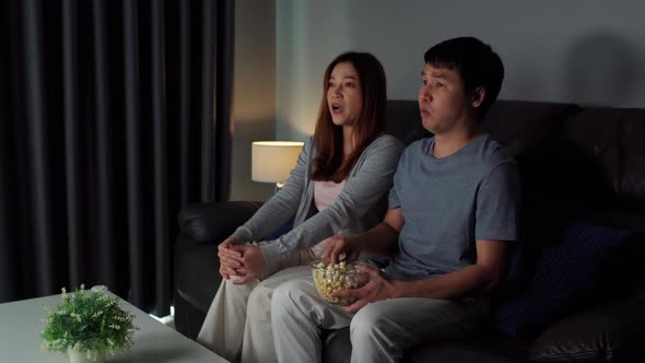 shocked young couple is watching horror movie TV on sofa at night