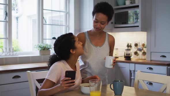 Mixed race lesbian couple hugging and drinking coffee in kitchen alt