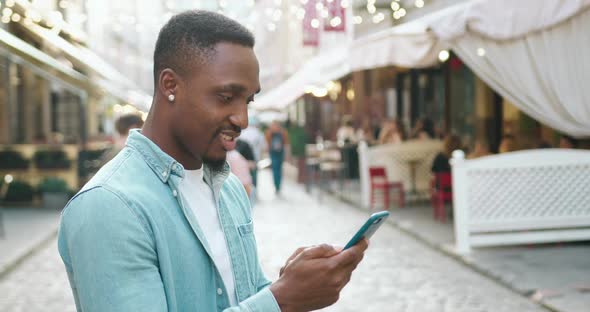 Black-Skinned Stylish Guy Standing on the Urban Street with Outdoors Cafes and Uses His Smartphone alt