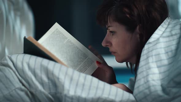Brunette Woman Covered with a Blanket Lies on the Bed and Reads a Book at Night Cinematic Shot