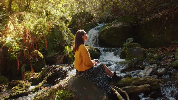 Young Woman is Sitting on the Rock Nearby the Cascading Water Stream Among the Autumn Forest