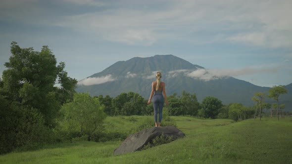 Spiritual yoga woman steps onto boulder raising arms into upward salute pose towards mount Agung alt