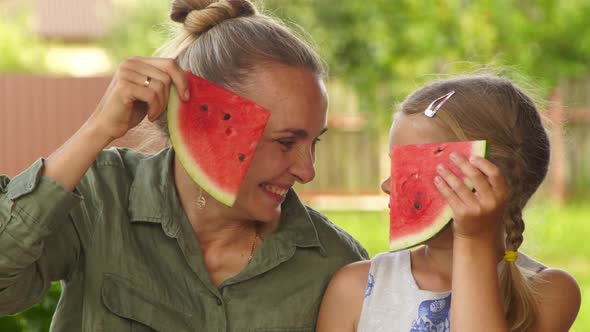 A Happy Mom and Cute Daughter Smiling and Hiding Behind Slices of Watermelon