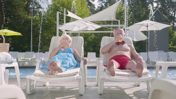 Mature Couple Lying on Sunbeds Near the Pool Drinking Juice, Talking and Smiling alt