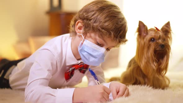 Charming Boy in Covid19 Face Mask Lying in Living Room Writing Letter to Santa Clause on Christmas alt