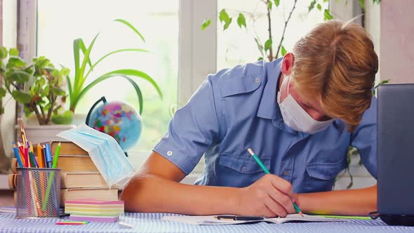 Teenage Man in a Blue Medical Mask of Caucasian Ethnicity Sits at His Desk at His Desk  alt