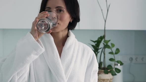 Thirsty smiling 35s woman in white bathrobe drinking water