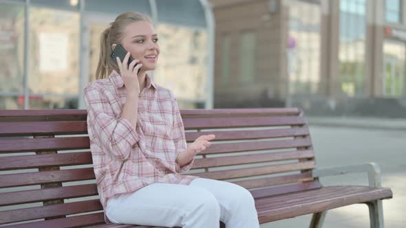 Young Woman Talking on Phone While Sitting Outdoor on Bench alt