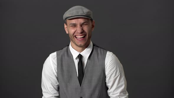 Portrait of Joyous Stylish Man Wearing Vest and Flat Kepi Laughing and Joking Over Gray Wall alt
