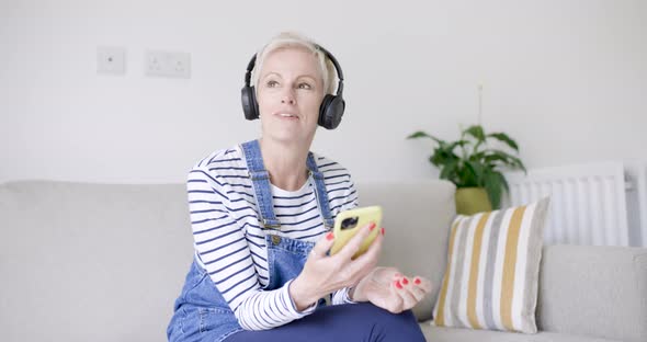 Mature woman sitting on couch listening music with headphones alt