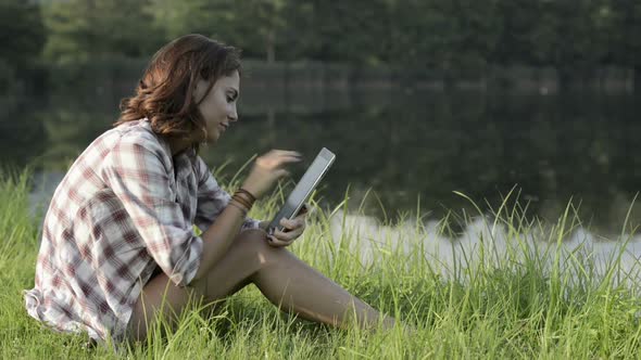 Young Woman in Nature Seated on Grass Look at Tablet on Lake Shore in Sunny Summer Day Outdoor