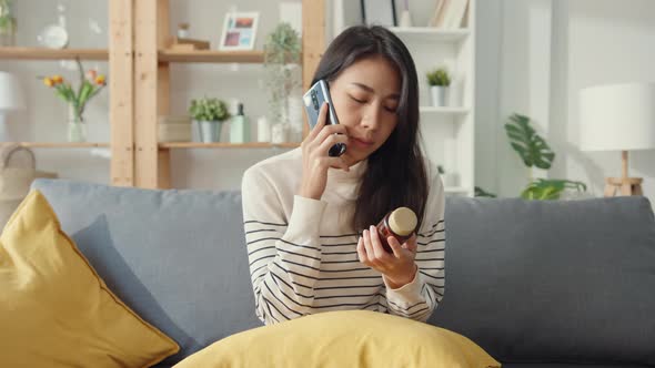 Asian woman hold medicine sit on couch use smartphone call to consult with doctor at home.