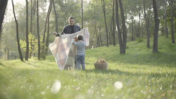 Wide Shot of Caucasian Young Father and Little Son Laying Blanket on Green Summer Meadow alt