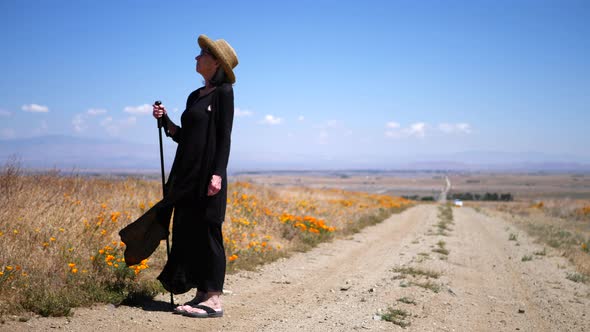An old woman in a dramatic black dress and sun hat on a dirt road under blue skies with flowers and alt