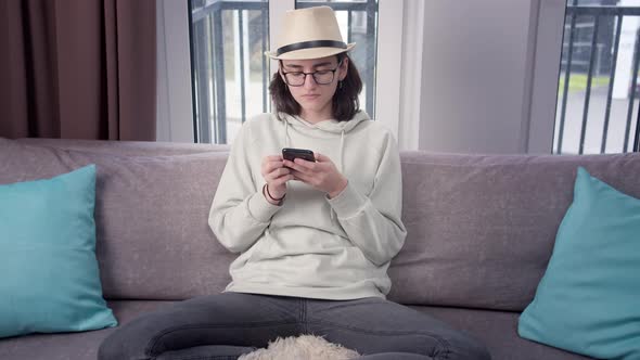 Brunette Girl in a Hat Uses Her Cell Phone at Home on the Couch Window Background alt