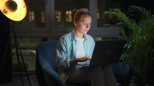Woman is Sitting in the Armchair and Working on a Laptop at Night alt