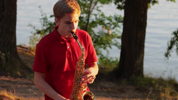 A young guy plays the saxophone near the river bank at sunset. Close-up.