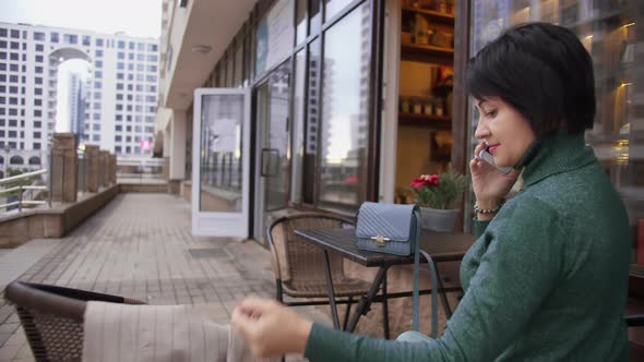 Woman with a Jacket Sitting in a Street Cafe and Calling on the Phone