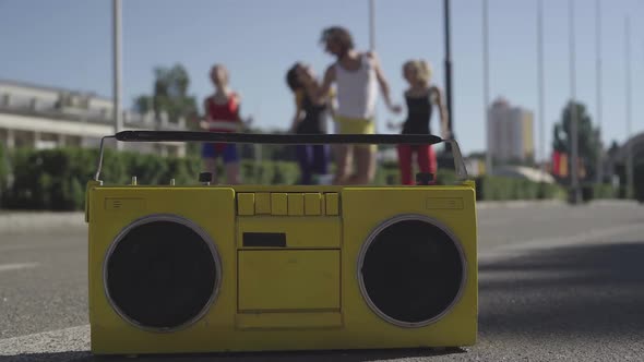 Retro Tape Recorder Standing on Asphalt with Four Caucasian Sportive People Doing Fitness Exercises alt