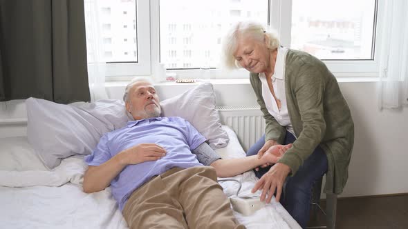 Caring Elderly Woman Measures Her Beloved Husband's Blood Pressure While Lying on a Bed in a Room alt