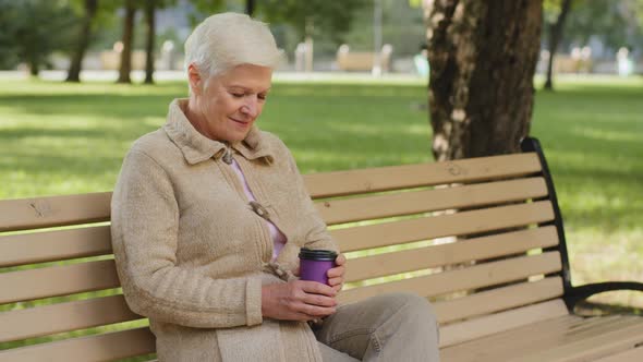 Dreamy Calm Relaxed Grayhaired Elderly Grandmother Sitting on Park Bench Drinking Coffee From Glass alt