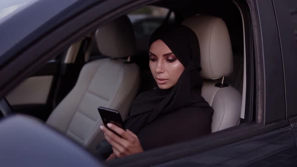 Attractive Young Woman in Black Hijab Sitting in Her Car Thoughtfully Looking at Her Smartphone alt