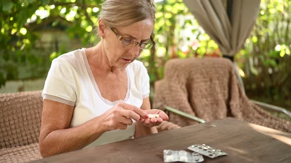 Elderly senior old woman in spectacles takes medicine drugs vitamins pills outdoors in the garden.