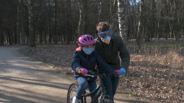 A Father Teaches His Daughter To Ride a Bicycle in a City Park. Protective Helmets and Medical Masks