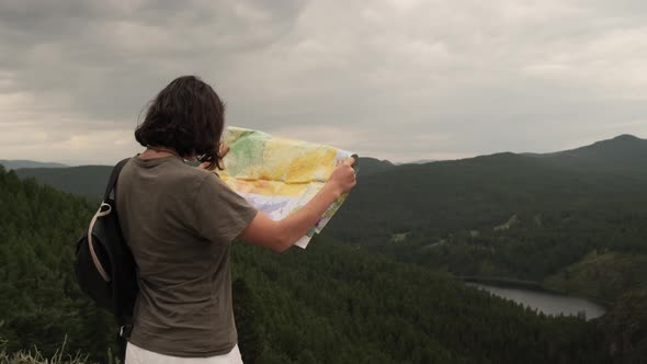 A young girl on a trip stands with a paper map against the background of mountains and a lake.