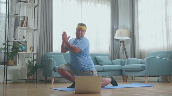 Fat Asian Man On Mat Looking At Laptop And Trying To Do Yoga In Law Lunge Pose At Home alt
