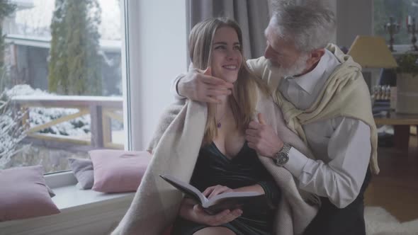 Middle-aged Caucasian Man Covering Reading Young Woman with Blanket. Cheerful Couple with Age Gap alt