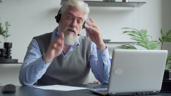 Smiling Elderly Man with a Gray Beard Wearing Headphones, Having a Video Call on a Laptop, Happy alt