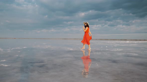 Young Woman Tourist in a Waving Dress Walks Along a Salt Lake in Which the Sky Is Reflected alt