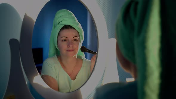Happy Woman Sitting in Front of a Mirror at Home and Applying Makeup with a Brush Reflection in the