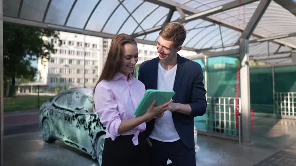 Positive Young Man and Woman Surfing Internet on Tablet Smiling Standing at Service Station with Car alt