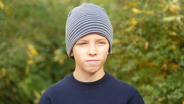 Serious Boy in Striped Hat Standing in Park Looks at Camera and Nods His Head in Agreement alt
