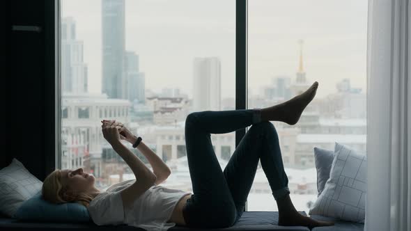 a woman with a phone lies by a large window overlooking the city