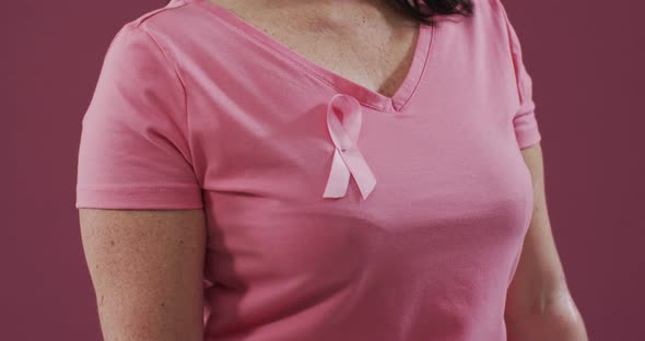 Mid section of a woman with pink ribbon on her chest clenching her fist against pink background alt